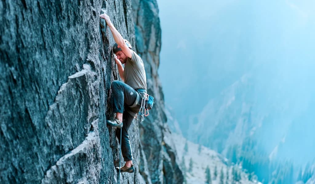 A climber reaches for a handhold on a steep granite rock face above a misty valley.