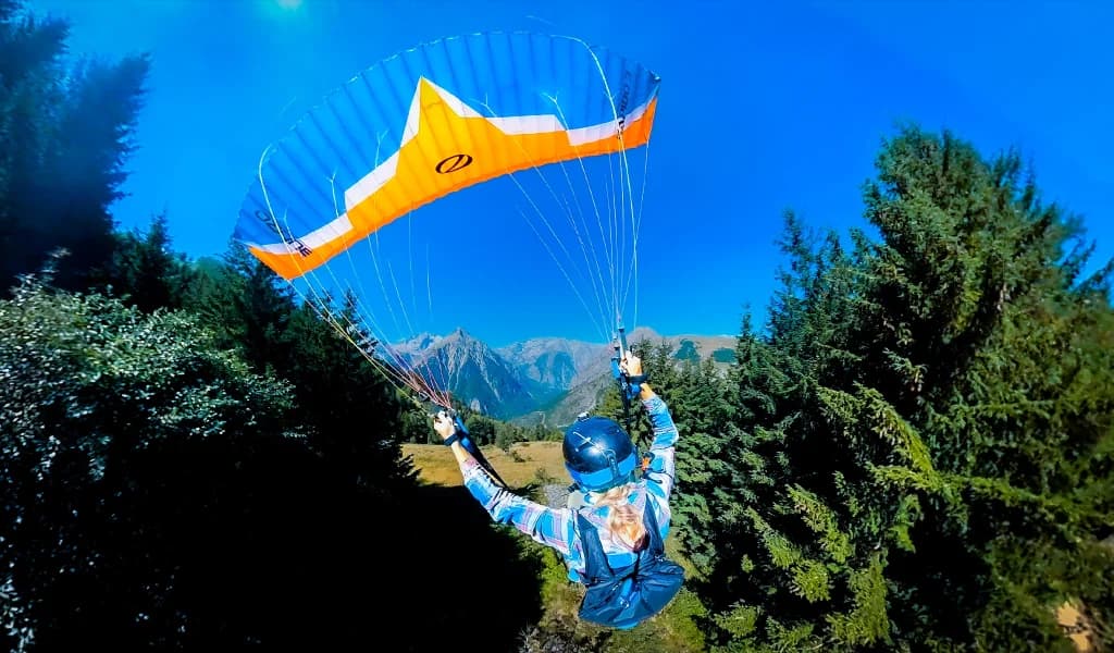 A paraglider in a colorful wing launches over green mountain trees under a clear blue sky.