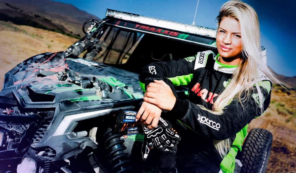 Christine GZ posing beside an off-road racing vehicle in a desert setting.