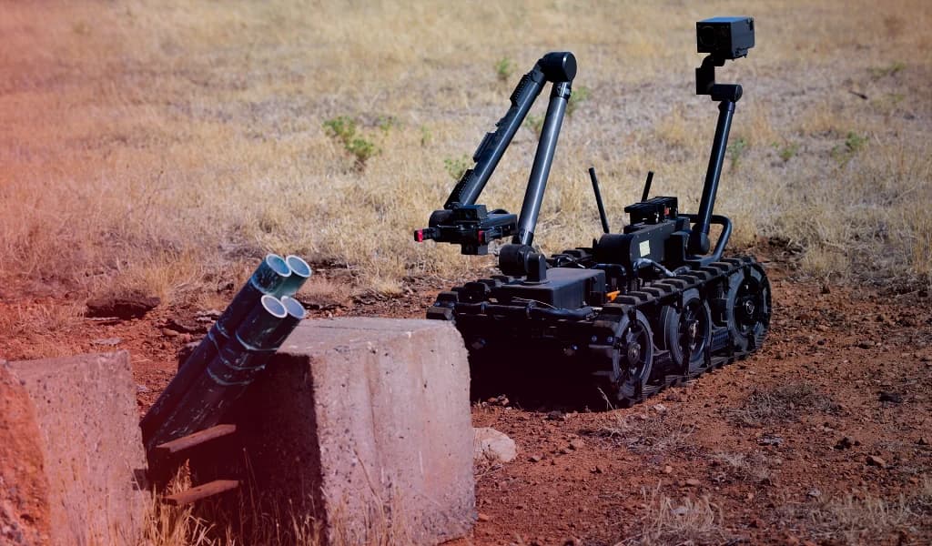 Tracked bomb disposal robot approaching a suspicious device in a dry, open landscape.