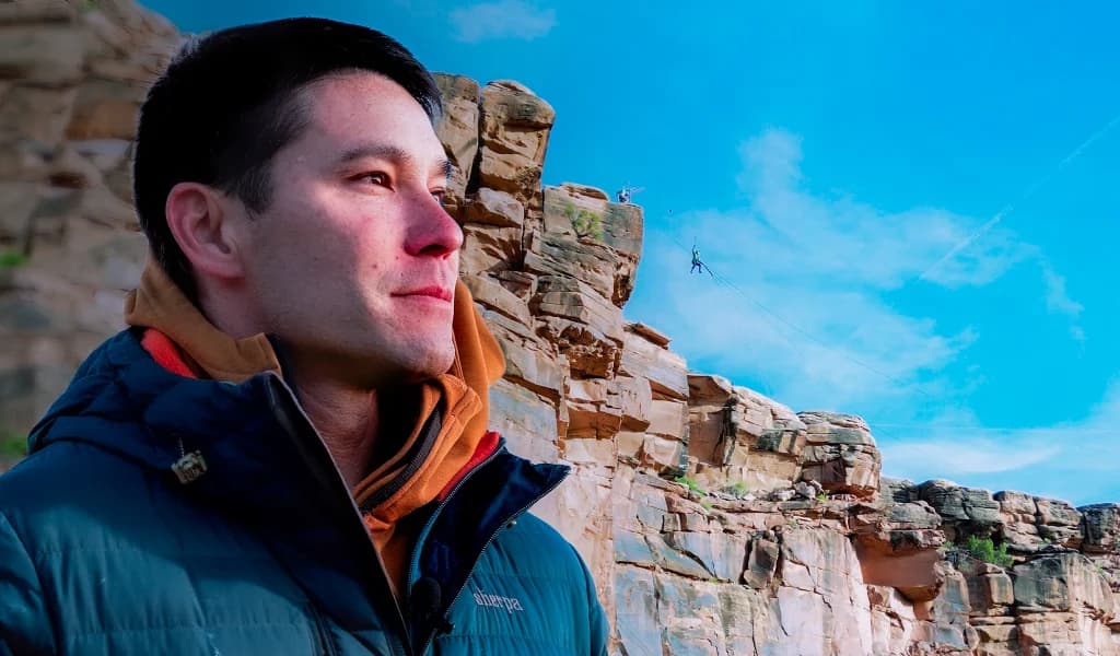 JJ Yosh looking toward the horizon with a person on a rope swing visible against red rock cliffs in the background