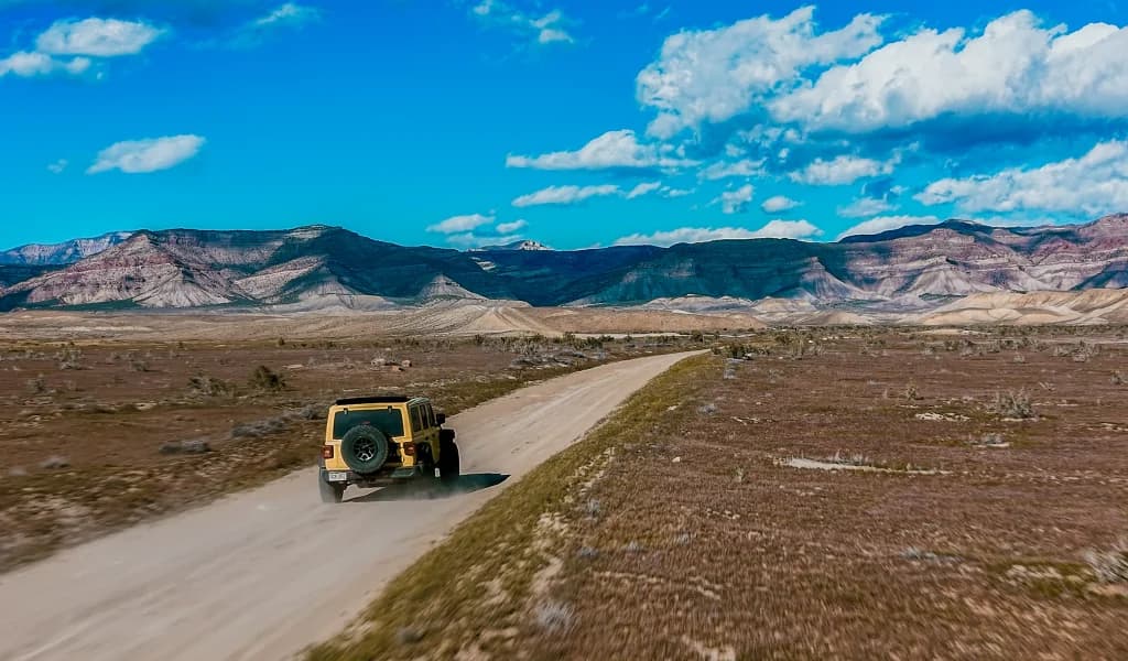 Yellow Jeep driving along a desert dirt road with layered mountain formations under a blue sky.