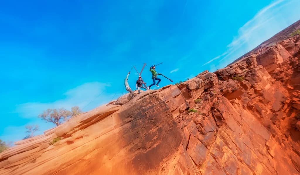 JJ Yosh leaping off a red rock cliff edge with a rigger and another person watching from the ledge above.