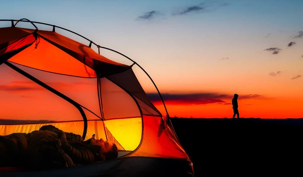 JJ Yosh lying in a glowing tent with a silhouette standing against a vivid desert sunset.