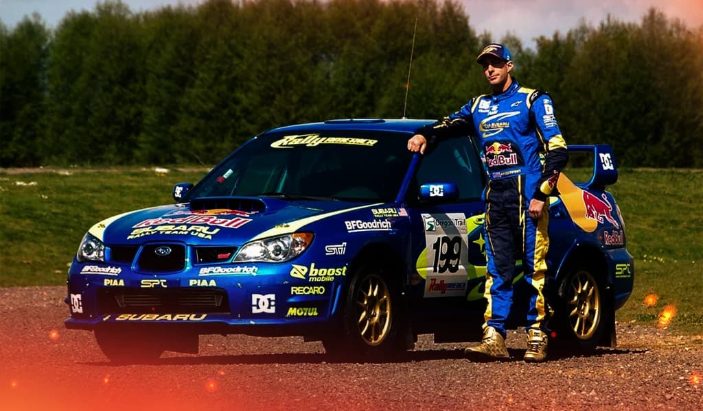 Racer stands beside a blue Subaru rally car with Red Bull and DC logos.