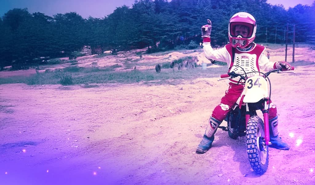 Young child, Travis Pastrana, in red motocross gear and helmet sitting on a small dirt bike, holding one hand up with a finger pointed in the air.