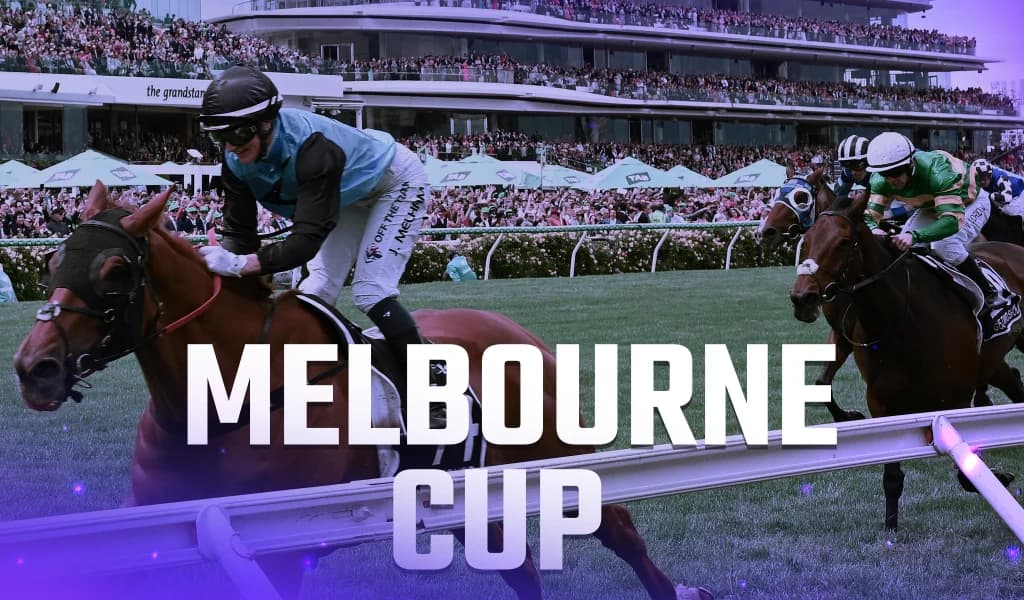 Jockeys riding racehorses down a grassy track with a large crowd in the grandstand.