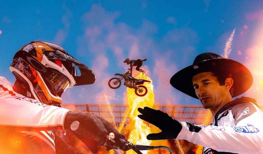 Two motocross riders, Selema Masekela and Jarryd McNeil, in the foreground with another rider jumping through flames during a freestyle stunt in the background.