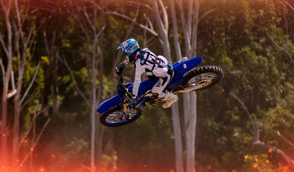 A motocross rider performs a sideways whip while airborne above a dirt track surrounded by trees.