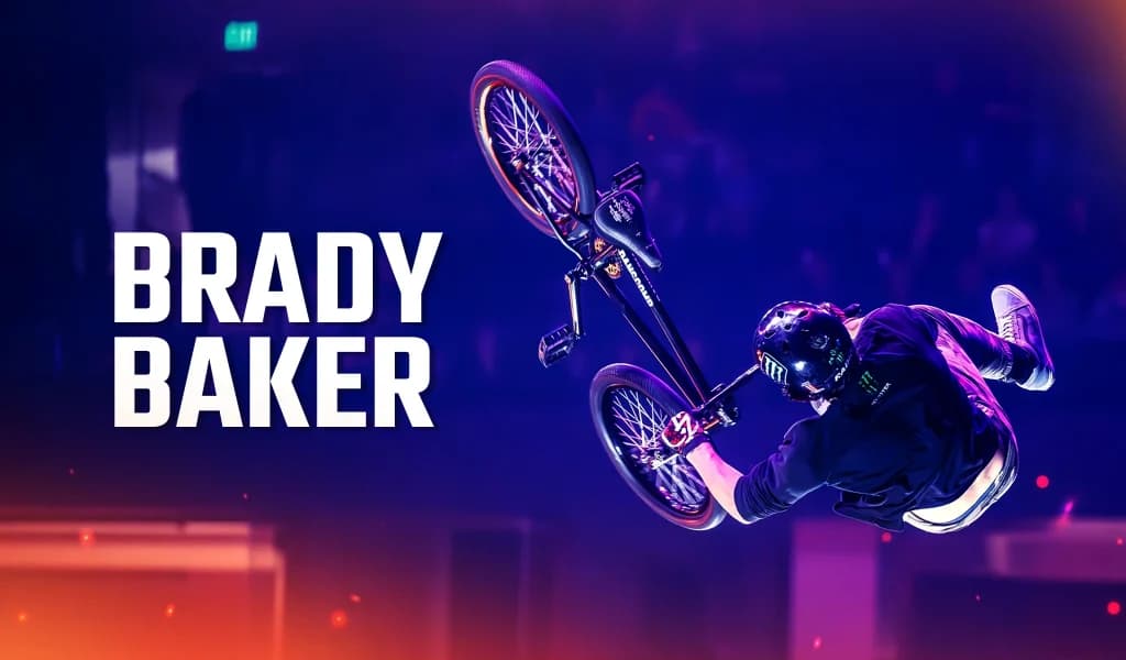 BMX rider Brady Baker rotating midair inside an arena with purple lighting and blurred crowd in the background.