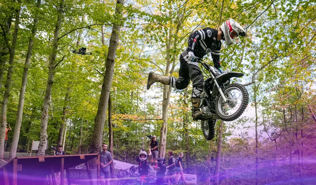 Pastranaland rider performing a freestyle jump on a pit bike in a forested area while spectators watch nearby.