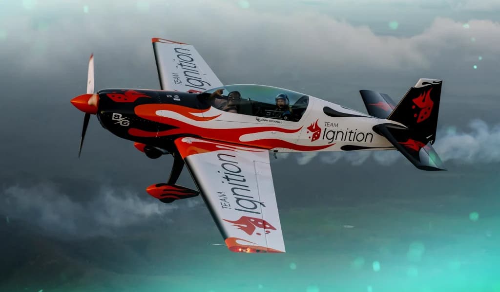 A small aerobatic plane wrapped in The Team Ignition Show branding flying above the clouds with two people inside the cockpit during an aerial maneuver.