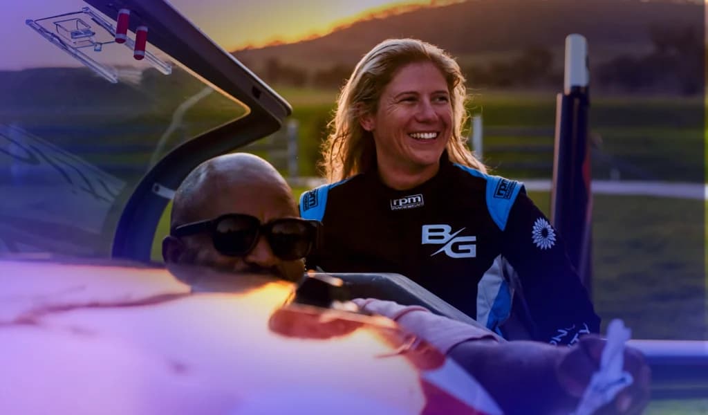 Two people smiling while seated inside the cockpit of a small aerobatic aircraft on the ground.