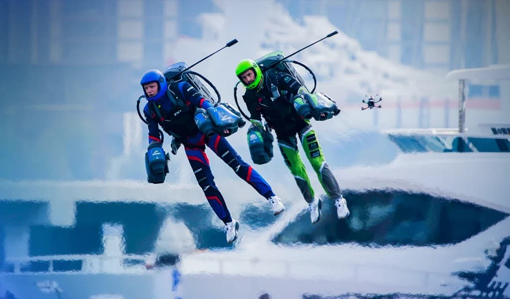 Two jet suit pilots flying side by side above water near boats, with a drone filming them.